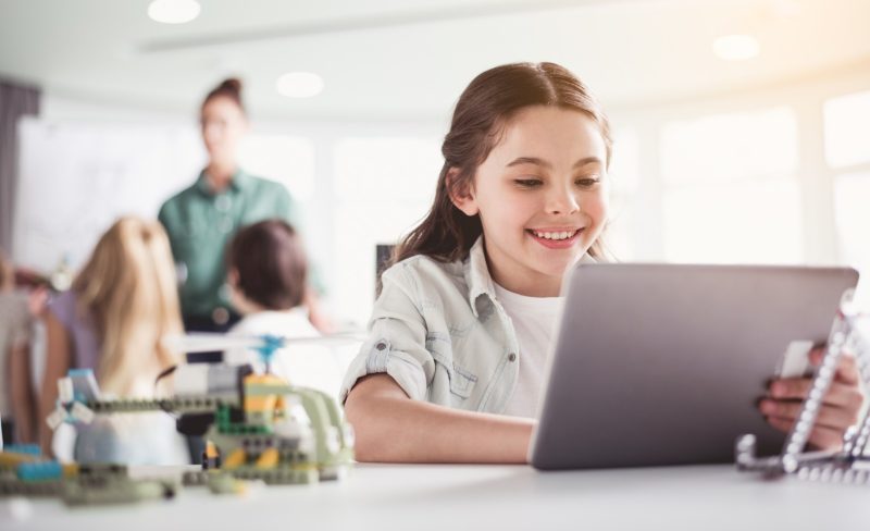 Portrait of cheerful child having fun with digital device while situating at table in classroom