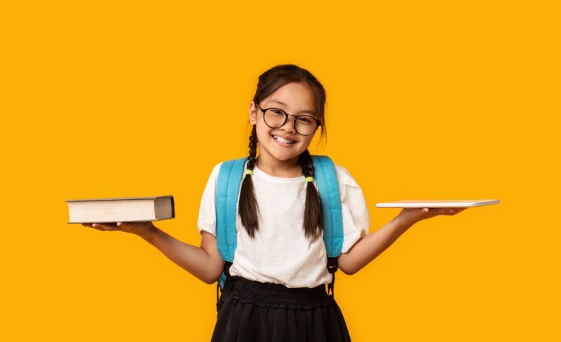 Gadgets Vs Books. Asian School Girl Comparing Digital Tablet And Book Standing Over Yellow Studio Background.